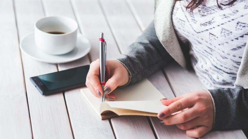 A person's hands writing in a small notebook with a pen on a white wooden table
