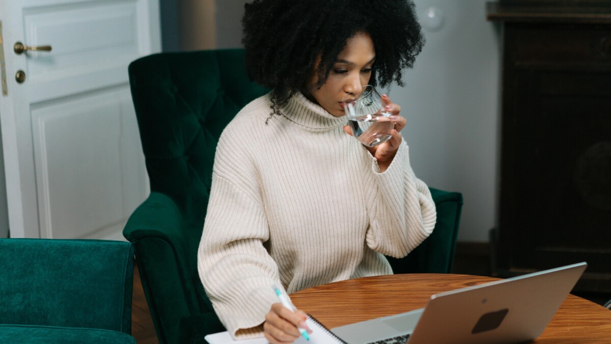 Person working remotely, focusing on her notes and computer, pausing to hydrate with a glass of clear water